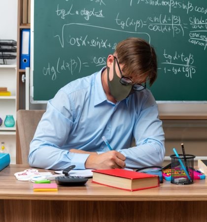 young-male-teacher-wearing-glasses-facial-protective-mask-preparing-lesson-looking-confident-sitting-school-desk-with-books-notes-front-blackboard-classroom_141793-94140